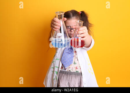 MR - Science Geek Young Girl / Female - Age 7 - holding borosilicate glass beakers of blue and red liquids out towards camera on yellow background Stock Photo