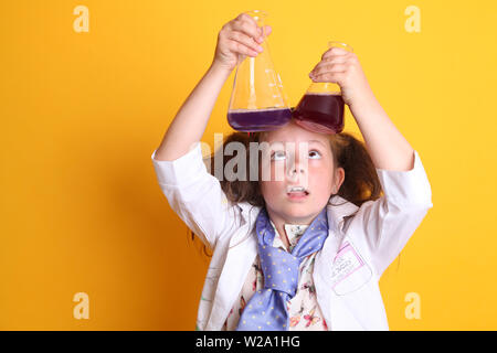 MR - Science Geek Young Girl / Female - Age 7 - looking up, holding borosilicate science beakers full of chemicals above her head on yellow background Stock Photo