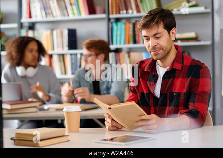 Handsome student with open book while sitting by desk in college library Stock Photo