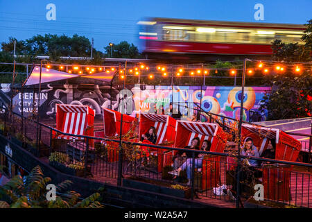 Young people in Else bar, a train, S-Bahn goes around, twilight, Alt Treptow, Berlin city life Germany, People sitting in wicker beach chairs Stock Photo