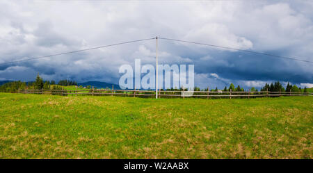 Electric power transmission pole in the mountains with cables along a wooden rail fence leading to the village. Panoramic landscape with a green field Stock Photo