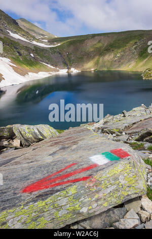 Bulgarian flag and red arrow track marking in front of the Eye lake, one of seven Rila lakes and distant waterfall and sunlit mountain peak Stock Photo