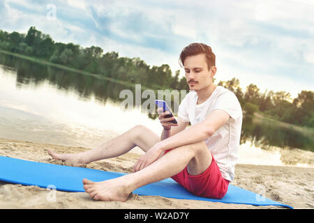 Man is sitting on the beach at lake and writing a sms on phone. good mood. beautiful view Stock Photo