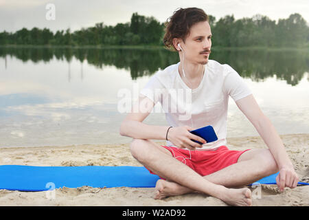 Young man walking near beautiful bay using phone and headphones for communicate with friends and listening music Stock Photo