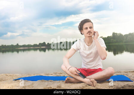 Young man walking near beautiful bay using phone and headphones for communicate with friends and listening music Stock Photo