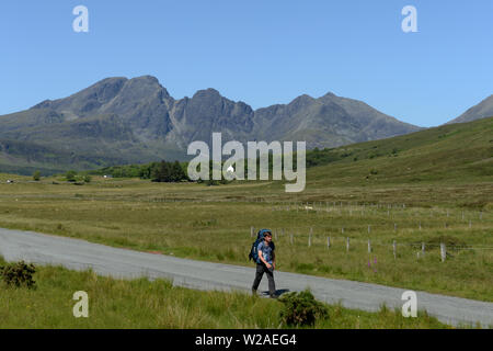 Back packer walking road to Elgol with Blà Bheinn or Blaven mountain on the Isle of Skye in Scotland and its distinctive jagged rocky outline visible. Stock Photo