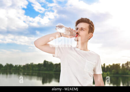 young guy drinks fresh water against of the lake and the forest background. summer thirst. beautiful view Stock Photo