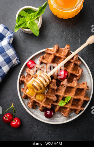 Whole wheat healthy belgian waffles with honey and cherries on plate, black concrete background, table top view sweet food or dessert