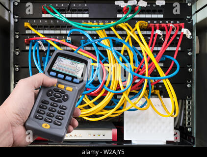 Network test. Qualified cable performance tester in human hand detail. Data signal measurement. Colorful cables connected in patch panels of rack case. Stock Photo