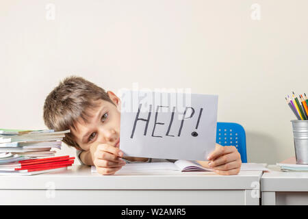 Sad tired frustrated boy sitting at the table with many books and holding paper with word Help. Learning difficulties, education concept. Stock Photo