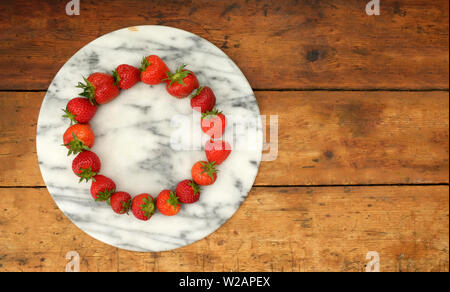 sixteen red ripe strawberries positioned in a circular shape on a marble circular board on a rustic wooden table top, still life with lots of copy spa Stock Photo