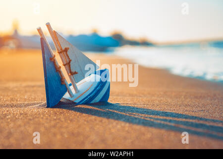wooden toy sailboat buried into sand near the seashore in summer. Vacation and leisure concept concept. Stock Photo