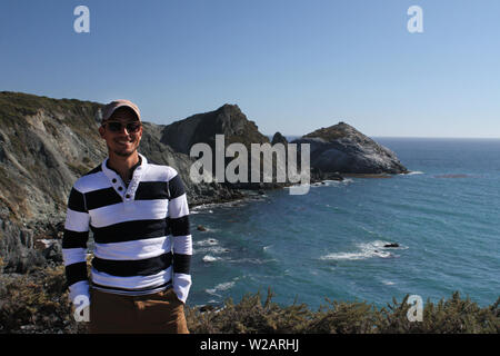 Smiling man wearing sunglasses standing next to a coastal panorama in California Stock Photo