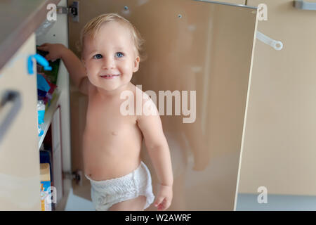 Cute curious caucasian baby boy open cupboard door in kitchen and exploring content. Funny toddler kid smiling and searching for sweets discovering Stock Photo