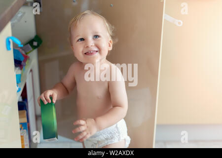 Cute curious caucasian baby boy open cupboard door in kitchen and exploring content. Funny toddler kid smiling and searching for sweets discovering Stock Photo