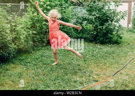 Preschool kids girl splashing with gardening hose sprinkler on backyard during summer day. Child playing with water outside at home yard. Candid authe Stock Photo