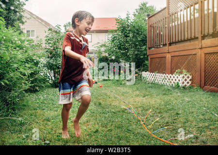 Preschool kid boy splashing with gardening hose sprinkler on backyard during summer day. Child playing with water outside at home yard. Candid authent Stock Photo