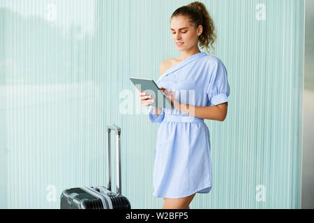 Positive attractive young woman standing at her suitcase and using tablet computer to figure out how to get to hotel Stock Photo
