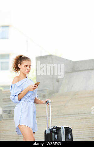 Serious young Caucasian woman standing in the street with suitcase and reading text message in her phone Stock Photo