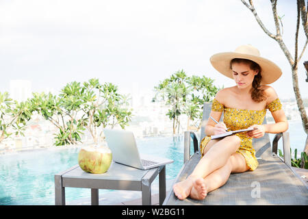Pretty young businessowoman sitting on chaise-lounge at swimming pool and taking notes in planner Stock Photo