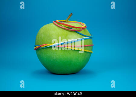 Green apple tied in colorful rubber bands over a blue background. Simplicity back to school or diet, aerobic concept with copy space for text Stock Photo