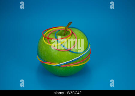 Green apple tied in colorful rubber bands over a blue background. Simplicity back to school or diet, aerobic concept with copy space for text Stock Photo