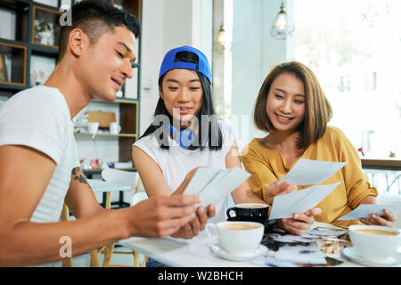 Group of positive young Asian people discussing printed photos when drinking coffee in cafe Stock Photo