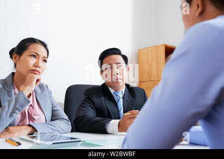 Concerned serious Vietnamese entreprneurs talking to young employee and discussing his mistakes Stock Photo
