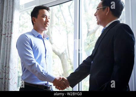 Cheerful young Asian businessman shaking hand of his mature coworker at meeting Stock Photo
