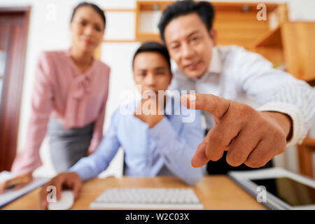 Mature Asian businessman pointing at computer screen of coworker, focus on index finger Stock Photo
