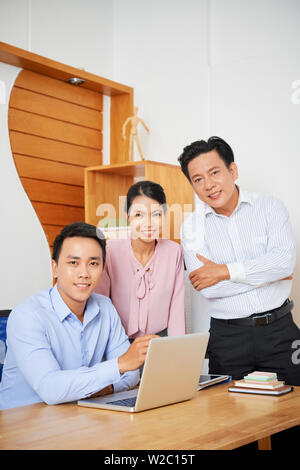 Team of cheerful Vietnamese business executives standing at office table of young smiling colleague Stock Photo