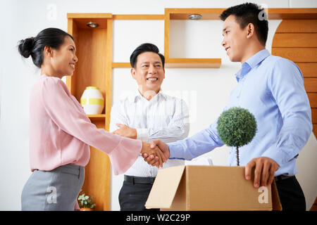 Young Asian businessman with cardboard box of belongings shaking hands of his new coworkers Stock Photo