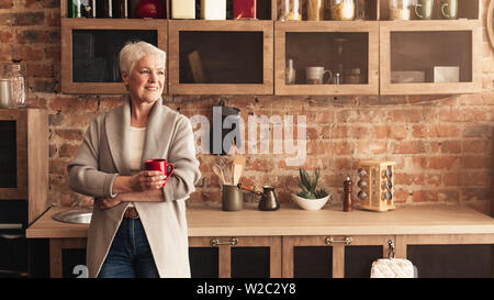 Calm senior lady standing at kitchen, drinking coffee Stock Photo