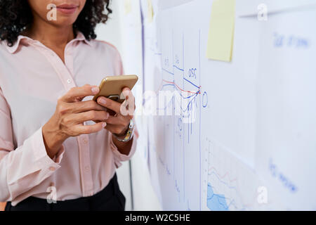 Businesswoman standing near wall with financial charts and reports and sending text message to her coworker Stock Photo
