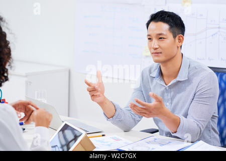 Smiling young Vietnamese entrepreneur sitting at table with financial charts and talking to female colleague Stock Photo