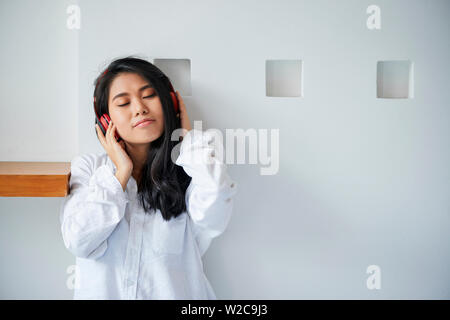 Beautiful Asian woman in white shirt wearing wireless headphones listening to music and enjoying it at home Stock Photo