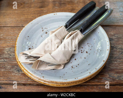 Two set of clean fork and knife wrapped in tissue napkin in the ceramic dish on wooden table. Stock Photo