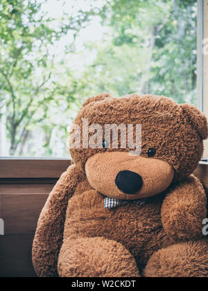 Big cute teddy bear with blue plaid bow tie sitting near the wooden window glass vertical style. Stock Photo
