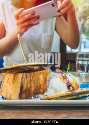 Woman's hands taking photo of the dessert, Ice cream with banana toast on wooden table by smartphone vertical style. Stock Photo
