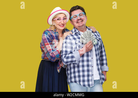 Happy wealthy family, adult man and woman in casual checkered shirt standing pickaback together, holding fan of dollar, toothy smile, looking at camer Stock Photo