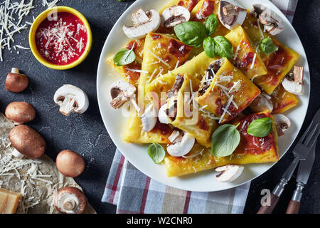 close-up of Crepes with parmesan and mushrooms filling on a white plate on a concrete table with tomato sauce, piece of cheese and kitchen towel, view Stock Photo