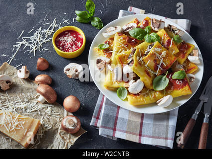 rolls of crepes with parmesan and mushrooms filling on a white plate on a concrete table with tomato sauce, piece of cheese and kitchen towel, view fr Stock Photo