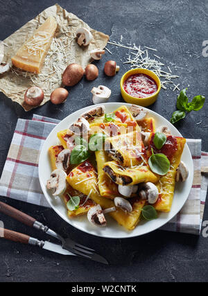 rolls of crepes with parmesan and mushrooms filling on a white plate on a concrete table with tomato sauce, piece of cheese, vertical view from above Stock Photo