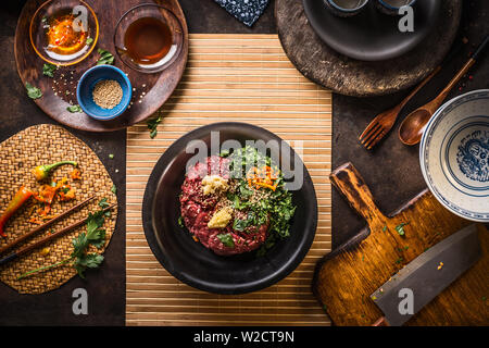 Mincemeat in black bowl with chopped garlic, chili, ginger and herbs on dark kitchen table background with sesame and soy sauce in bowls, top view. St Stock Photo