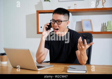 Asian young businessman in black T-shirt sitting at his workplace in front of laptop talking on mobile phone and angry Stock Photo