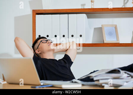 Young Asian businessman in eyeglasses and in casual clothing relaxing with eyes closed on chair opposite his workplace he tired of work Stock Photo