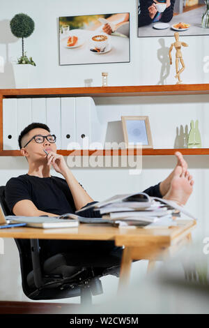 Asian young office worker in eyeglasses sitting in comfortable pose at his workplace and thinking about new ideas while working at office Stock Photo