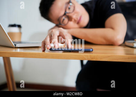 Asian office worker lying on his workplace and touching the display of his smartphone while working at office Stock Photo