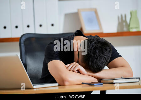 Young businessman in casual black T-shirt sleeping at his workplace with laptop and notepad during work day at office Stock Photo