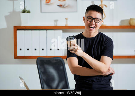 Portrait of Asian casual businessman in eyeglasses drinking coffee from disposable cup and laughing while standing near his workplace at office Stock Photo